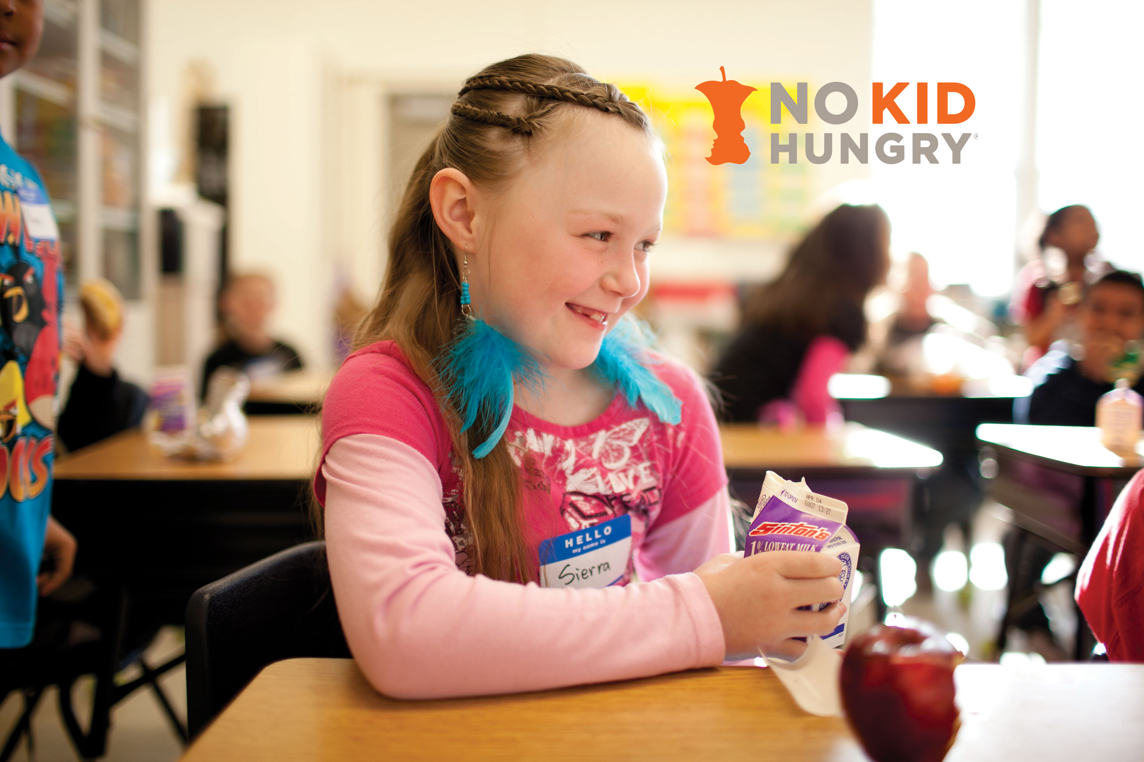 Smiling child in classroom holding a milk with an apple on her desk