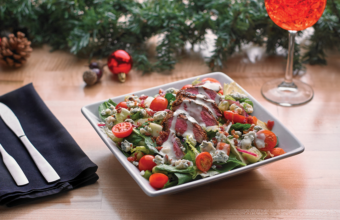 steak salad with tomatoes on a white bowl sitting on a wood table. 